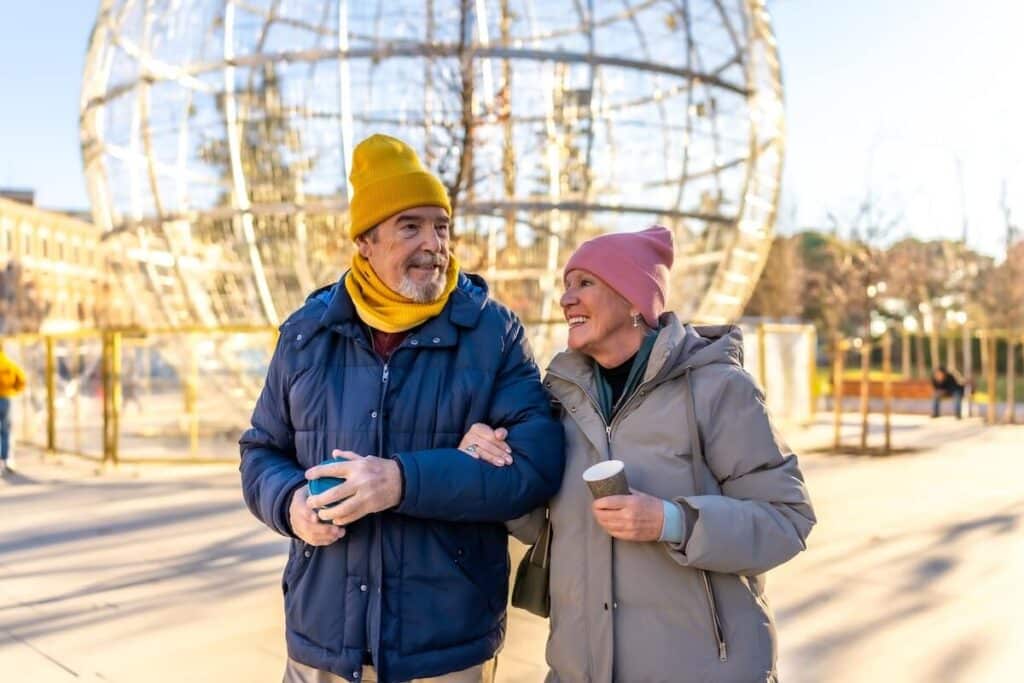 Smiling senior couple walking arm in arm in a winter city park with a large christmas decoration and disposable coffee cups in their hands